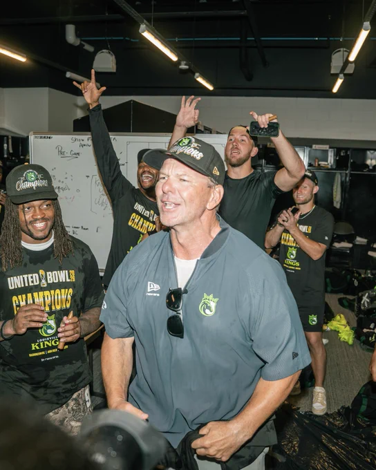 A jubilant sports team celebrates in their locker room after winning the United Bowl championship. The atmosphere is lively as team members wear championship shirts and hats.