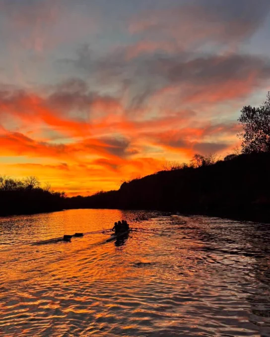 Rowers glide across the Iowa River at sunset with vibrant orange and pink clouds reflecting on the water.