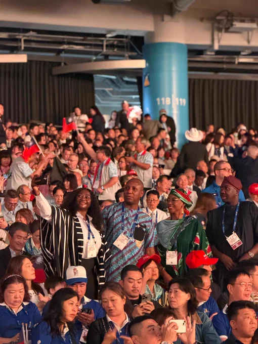 A diverse group of Rotary convention attendees stands together in a crowded arena, taking a selfie before the opening session. Participants wear colorful traditional and professional attire, representing different countries and cultures, while hundreds of attendees fill the seats behind them.