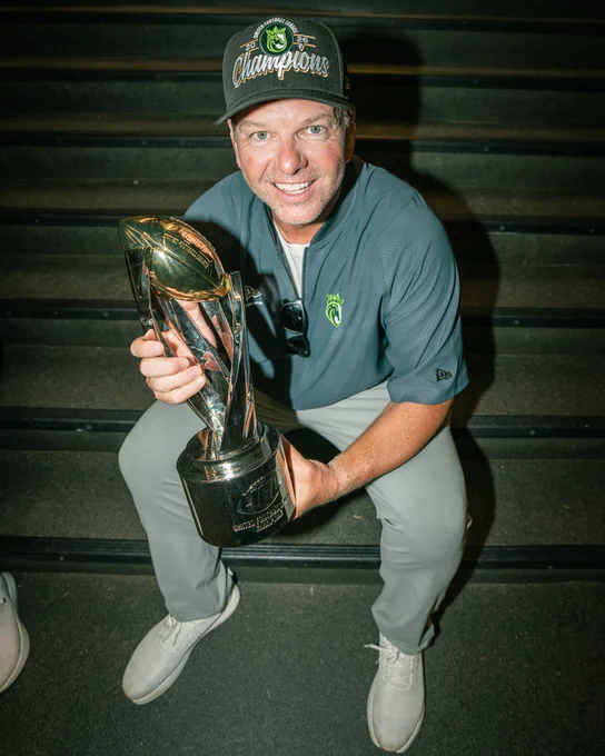 A man wearing a 'Champions' hat sits on stairs holding a shiny championship trophy. He appears happy and triumphant.