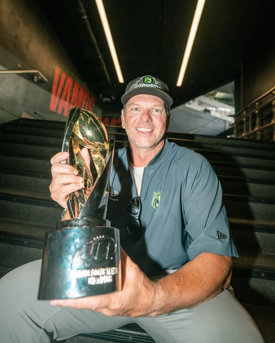 A man sits on stadium steps proudly displaying a shiny trophy. The setting is energetic, capturing a sense of achievement.
