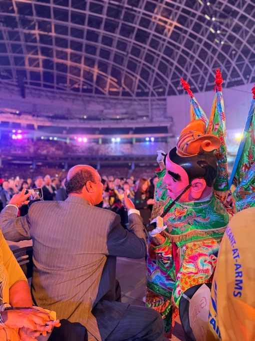 Traditional dancers and drummers perform on stage beneath colorful lighting during the opening ceremony of the Rotary International Convention. Temple-inspired imagery fills the large screen behind them as the audience watches from the arena floor and stands.
