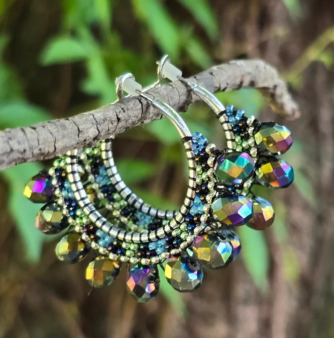 silvertone hoop earrings with beads in shades of iridescent black, purple, and green, with faceted iridescent beads, background of green leaves and brown tree bark