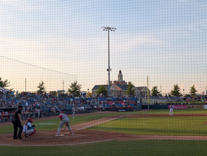Photograph of a baseball field from behind the home team dugout. The away team is at bat to the left with the umpire and catcher visible behind them. The third baseman is visible to the right. Behind the field is the town hall and a Carolina sunset.