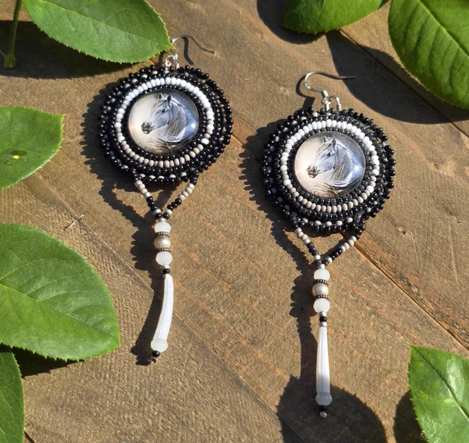 beaded earrings with round centers featuring a white horse head, surrounded by beads in shades of white, gray, and black, with dentalium shells, freshwater pearls, and silver ear wires, on a background of natural wood and green rose leaves
