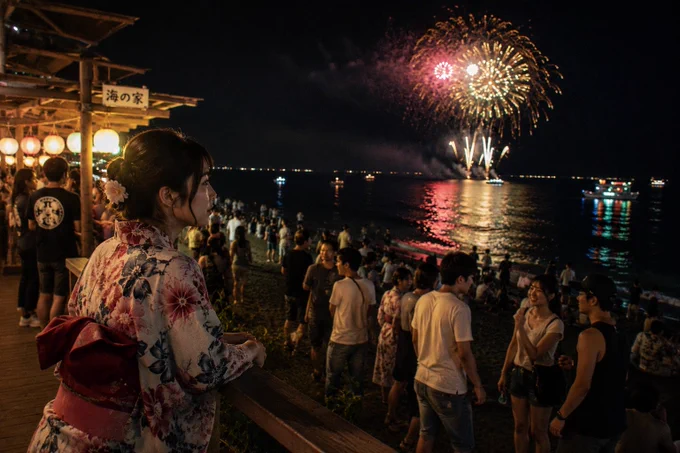 1枚。海辺の花火大会の賑わいを見下ろす流れ。

木製の階段や手すりが続く海沿いの施設から砂浜を見下ろしている。左側には複数階の海の家が並び、提灯が吊られたテラス席に人が集まっている。木材には木目や節が残り、提灯の紙は内側の灯りで透けて見える。

砂浜には大勢の人が集まり、浴衣姿の人、半袖姿の人、帽子をかぶった人が混ざって海の方を見たり立ち話をしたりしている。波打ち際まで人影が続き、小さなグループごとに立ち止まっている。

さらに奥では数隻の船が海に浮かび、白や緑の灯りが揺れる水面へ細長く映り込んでいる。夜空には大きな花火が開き、放射状に広がる光が海面にも反射している。花火の明るさと海辺の灯りが重なりながら、人の流れと海の景色が一続きになっている。

右下に焼き付けクレジットあり
created and described with AI assistance