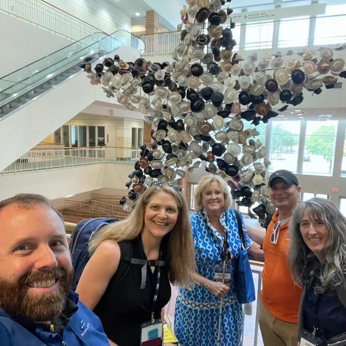 Five adults pose on an upper-level balcony inside a bright convention center. From left to right: a man, a woman, a woman, a man, and a woman. Behind them, a large hanging star-shaped display is covered in cowboy hats.