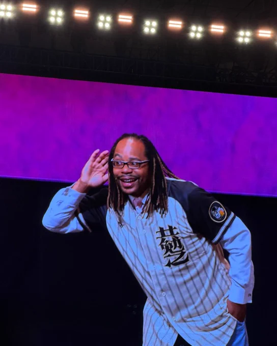 Jason Browne wears a baseball jersey over a dress shirt and listens to the audience with a hand cupped to his ear. He stands on stage beneath bright lights during the Rotary International Convention opening session.
