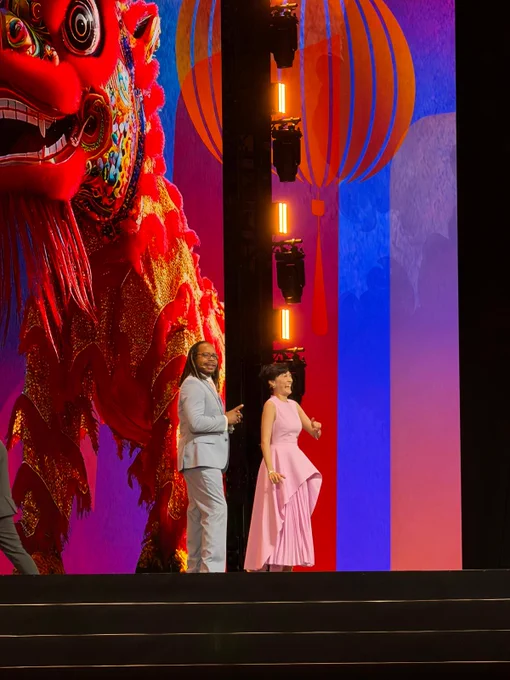 Betty Pei-hsin Chen and Jason Browne walk across the convention stage. Behind them, a colorful screen displays a traditional lion dance figure and hot-air balloons, celebrating Taiwanese culture during the Rotary International Convention opening session.