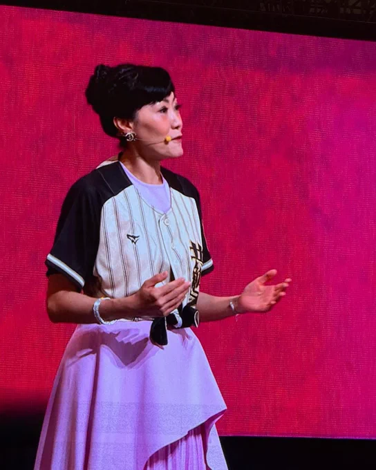 Betty Pei-hsin Chen stands on stage wearing a baseball jersey over a pink dress and a headset microphone. She gestures while speaking in front of a bright pink screen during the Rotary International Convention opening session.