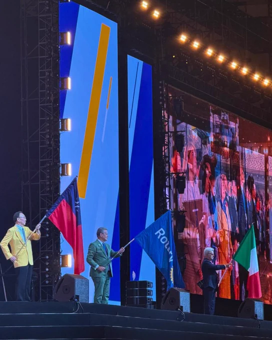 Three people stand on a brightly lit convention stage holding the flags of Taiwan, Rotary, and Italy during a flag ceremony. Large LED screens and stage lighting tower behind them as attendees appear on a massive screen in the background.
