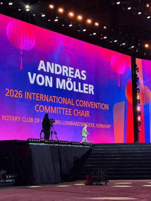 A giant convention screen displays “Andreas von Möller, 2026 International Convention Committee Chair” as he walks across the stage. Pink and blue graphics with lantern illustrations fill the backdrop, while a camera operator films from the side of the stage.