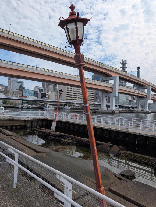 阪神・淡路大震災の被害をそのまま残した「神戸港震災メモリアルパーク」の風景。手前には大きく傾いた赤茶色のレトロな街灯が立ち、その奥の崩壊した岸壁や傾いたもう1本の街灯が痛々しい震災の爪痕を伝えています。背景には、復興を象徴するように綺麗に再建された2段の高架道路（阪神高速道路）や、近代的なビル群、そして白い雲が広がる青空が見えます。