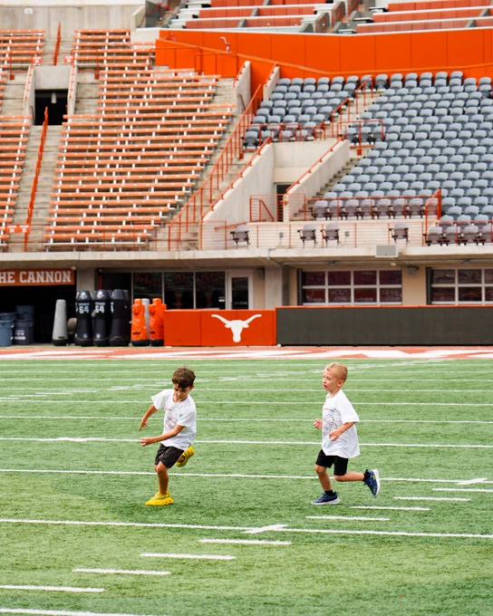 Two young boys in white t-shirts and shorts run on a green football field. Behind them are orange and gray stadium seats. A white Longhorn logo is on a wall.
