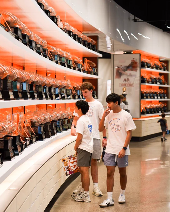 Three young men in white \"Dream. Speak. Live.\" t-shirts admire a vast, curved display of clear trophies on brightly lit orange shelves. A child is in the distant background.