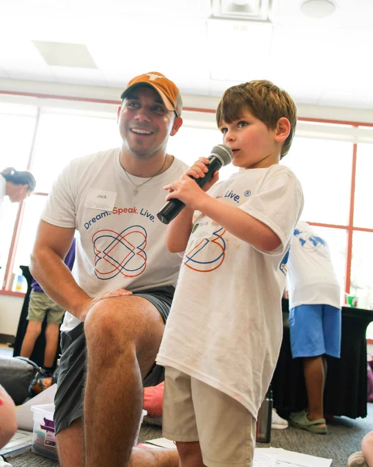 A smiling man, Jack, in an orange cap and white \"Dream. Speak. Live.\" shirt, kneels beside a young boy. The boy, also in a white shirt, speaks intently into a microphone.