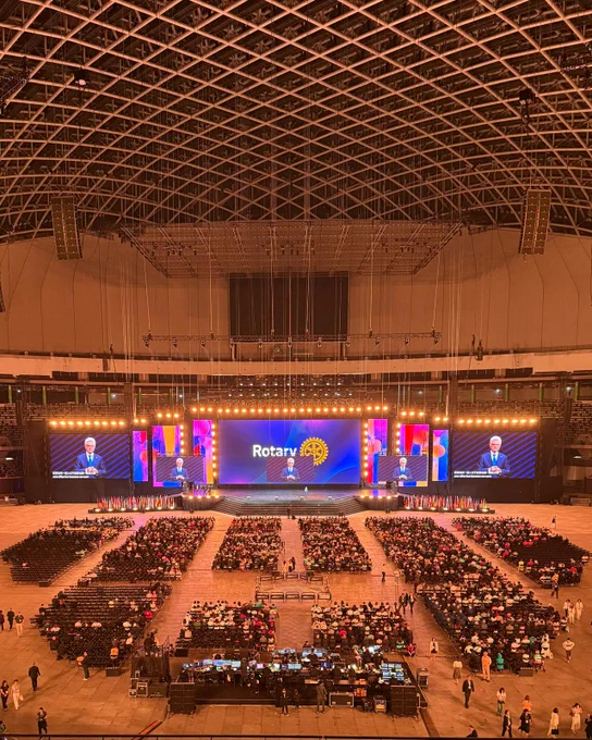 A wide view of Taipei Dome shows Rotary International President Francesco Arezzo speaking to thousands of attendees during the Opening General Session at #Rotary26. Large screens display his image while rows of international flags line the front of the stage.