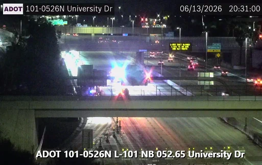 Traffic scene at night on a highway with flashing emergency vehicle lights near University Drive. The image shows a closed lane and a visible overhead electronic sign. Several other vehicles are near the scene, and an overpass is in the foreground. The date and time stamp reads 06/13/2026, 20:31:00.