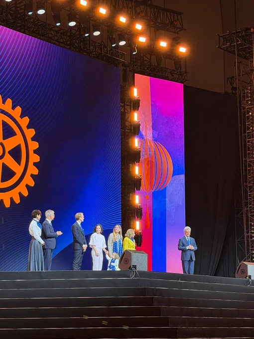 Rotary International President Francesco Arezzo stands on stage with several family members and Rotary leaders during the Opening General Session at #Rotary26. Colorful lantern graphics and the Rotary wheel logo appear on large screens behind them.