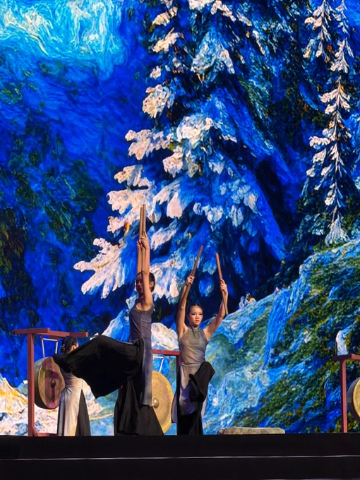 Performers raise wooden mallets during a choreographed percussion performance on stage. A towering digital backdrop depicts a snowy forest landscape in rich blue and white tones.