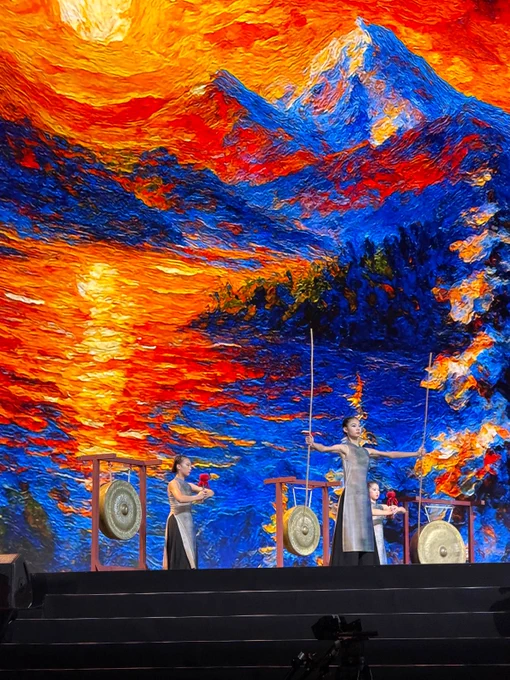 Performers stand on stage beside traditional gongs during a cultural performance. A vivid backdrop of mountains, water, and a glowing sunset fills the giant screen behind them in bright shades of orange, red, and blue.