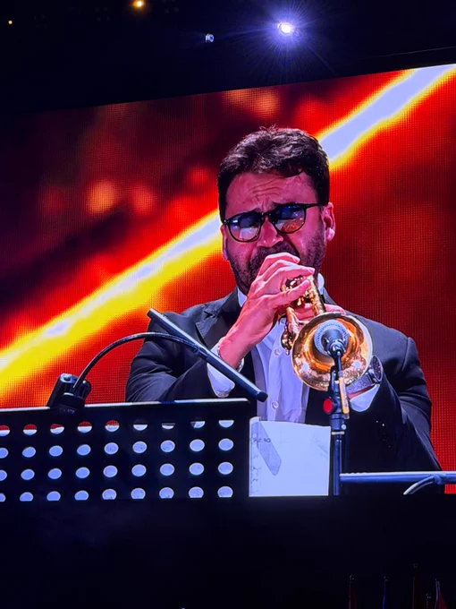 A trumpet player performs a solo during the HJO Jazz Orchestra's concert. Warm red and orange stage graphics illuminate the background as the musician plays under a spotlight.