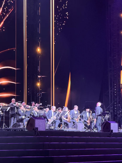 The HJO Jazz Orchestra performs on stage as musicians play brass and woodwind instruments under concert lighting. A conductor stands at the front of the ensemble against a backdrop of glowing gold accents.