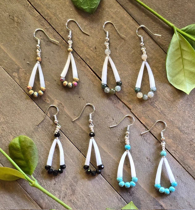 earrings made with 2 white dentalium shells each, with beads in shades of iridescent, turquoise, black, and green jasper with silver-tone ear wires, on a background of natural wood and green jasmine leaves 