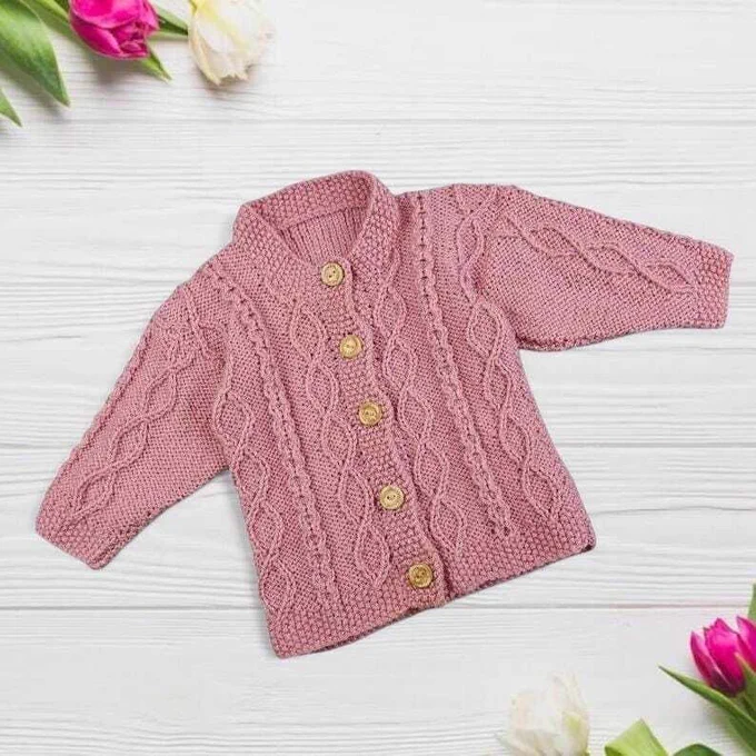 A dusty pink, cable-knit baby cardigan with five wooden buttons lies flat on a white wooden surface. Pink and white tulips decorate the upper-left and lower-right corners, creating a bright, clean aesthetic.
