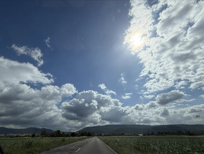 Das Foto zeigt eine lange, gerade Landstraße, die mittig durch eine weite ländliche Landschaft führt. Die Straße verläuft auf den Horizont zu und erzeugt eine starke Tiefenwirkung. Links und rechts der Fahrbahn erstrecken sich grüne Felder und Wiesen.

Im Hintergrund erhebt sich eine sanfte Hügel- bzw. Bergkette, die die Landschaft begrenzt. Vereinzelt sind Gebäude und Bäume zu erkennen, die auf kleine Ortschaften oder landwirtschaftliche Anwesen hindeuten.

Den größten Teil des Bildes nimmt der Himmel ein. Er präsentiert sich in einem kräftigen Blau und wird von zahlreichen großen, weißen bis grauen Quellwolken durchzogen. Die Sonne befindet sich im oberen rechten Bildbereich und scheint teilweise durch die Wolken hindurch. 

Die Stimmung des Bildes wirkt weit, ruhig und naturverbunden. Gleichzeitig verleihen die dramatischen Wolkenformationen und das intensive Sonnenlicht der Szene eine beeindruckende und dynamische Atmosphäre.