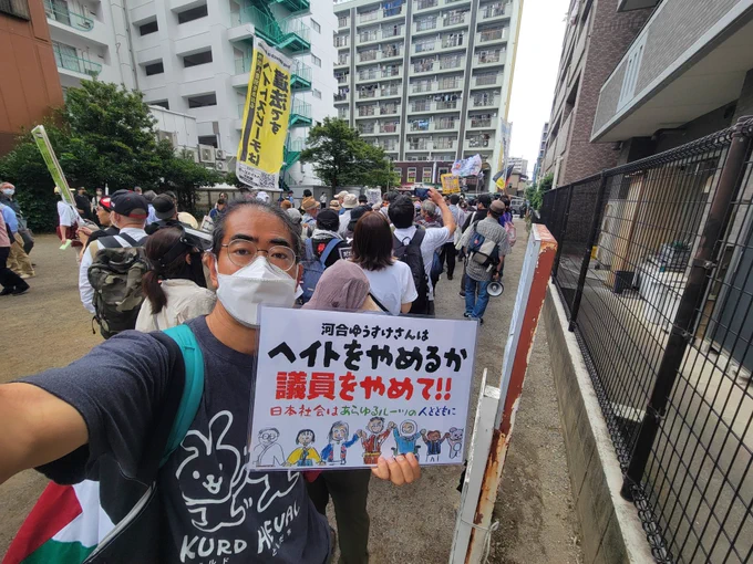 『河合ゆうすけさんはヘイトをやめるか議員をやめて!! 日本社会はあらゆるルーツの人とともに』と書かれた様々な属性の人たちが笑顔で手を繋いでいるクレヨン画のイラスト入りプラカードを持ってデモの待機列をバックにセルフィー