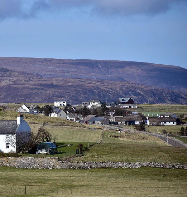 Today's 'Where in Scotland' image. It is a squarish image showing a view over fields to a settlement of one or two-storey dwellings, mainly finished in white. A road comes in the right edge of the frame and continues across part of the middle. There is a visible edge across the middle of the frame with, beyond it, a distant dark hillside climbing to the horizon. The scene is in sunlight.