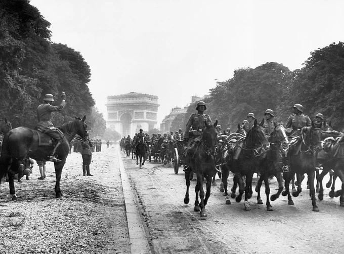 Historical photograph of soldiers on horseback riding down a broad, tree-lined avenue. The Arc de Triomphe is visible in the background. A large group of soldiers is proceeding along the road, with some onlookers at the sides.