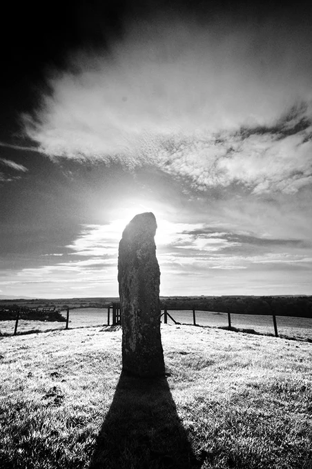 Atmospheric black and white photo of a large standing stone, back-lit by the sun.