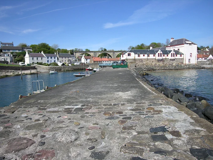 Lower Largo. The image shows a view along a broad stone pier to a village at its landward end. There is a large beige building on the right and houses on the left, with three arches of a railway viaduct visible in the centre of the frame. The sky is largely blue.