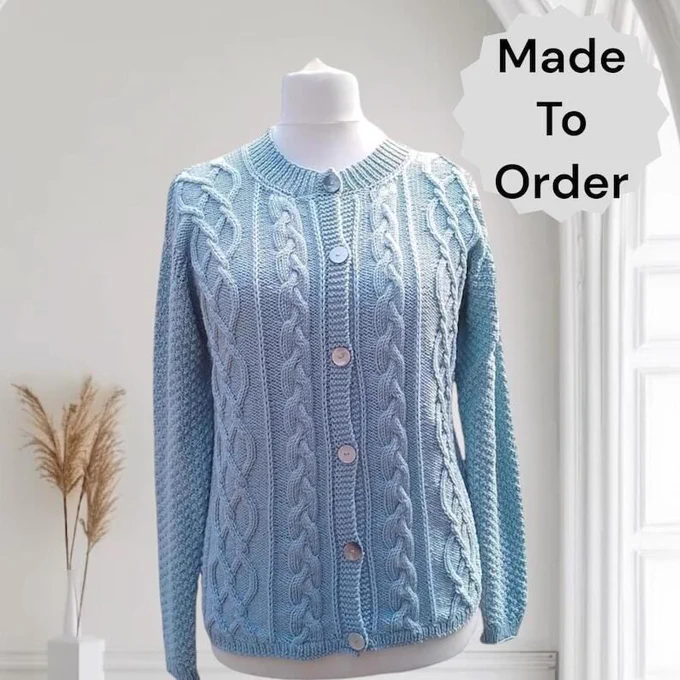 A pale blue knitted cardigan with intricate cable and textured stitching, round neck, and light buttons down the front, displayed on a white mannequin torso. Background: plain light wall, white trim/door frame to right, white vase with tall dried grasses left. Text overlay: “Made To Order” in black on white starburst shape.