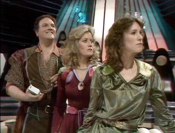 GPT 5.4 Mini describes the image as: "A scene from the BBC sci-fi series Blake’s 7, set on a futuristic interior with angular metallic walls, glowing panels, and a sleek, theatrical control-room feel. Three characters stand together, tense and alert, as if observing or reacting to events offscreen. The setting suggests an advanced spacecraft or base environment. David Jackson appears at left, holding a small device and looking upward with a slight grin; Sally Knyvette is centered, wearing a maroon outfit and looking on with concern; Jan Chappell is at right, in a shiny green costume, facing forward with a focused, determined expression. The image captures the show’s distinctive blend of adventure, sci-fi, and stylized 1970s production design."
The script search url for this tweet is
https://blake.torpidity.net/s/105/503