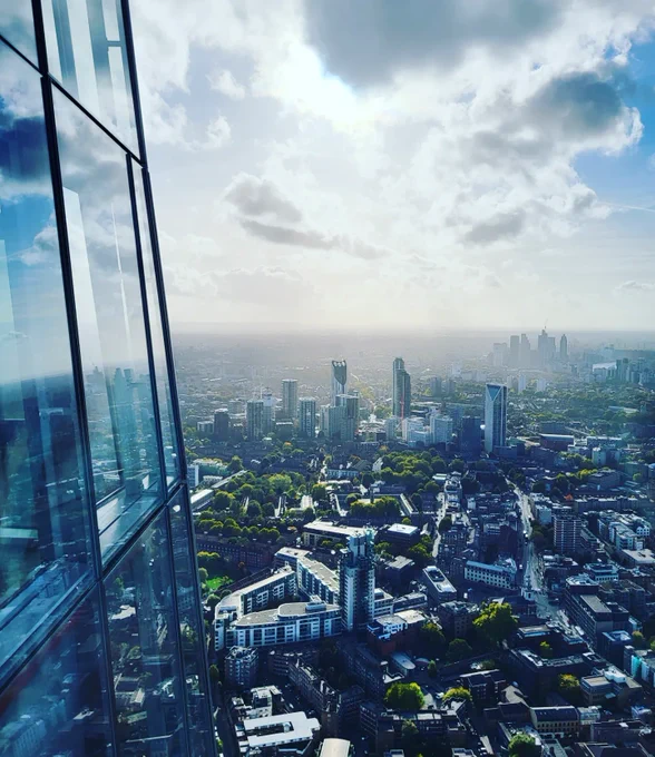 Aussicht vom Hochhaus the Shard in London. Links im Bild sieht man einen kleinen Teil der Glasfassade des Turms, in der sich Wolken und andere Gebäude spiegeln. Darüber hinaus sieht man einen Teil Londons mit anderen Hochhäusern, Bäumen und Straßen. Am Himmel sind Wolken zu sehen. Die Sonne versteckt sich hinter den Wolken und schafft es dennoch, den Himmel hell zu erleuchten.