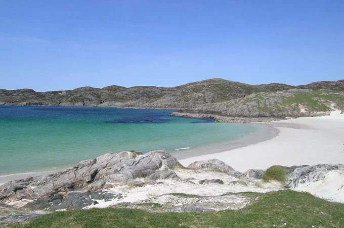 The image shows the beach at Achmelvich in the middle vertical third of the frame, with bright white sand on the right extending into light blue and then dark blue sea on the left. There are two tiny people walking on the beach. In the foreground the beach ends with rounded grey rocks, and there is sandy grass at the foot of the frame. Beyond the beach is a grey rock headland that cuts across the frame. The sky is blue. The front cover of ‘A Tangled Web’ is shown in the top right corner.