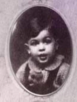 A portrait of a young boy with short hair, partially open mouth, and a collared garment.