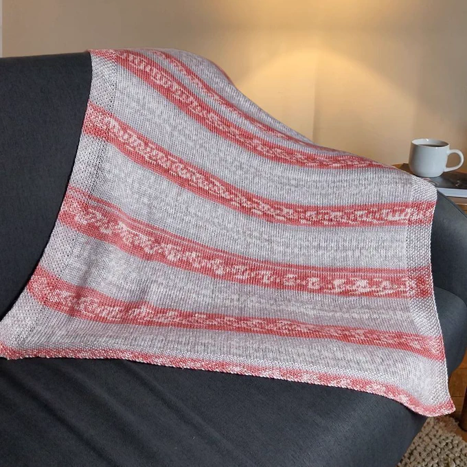 A knitted blanket with light beige and salmon pink horizontal stripes draped over a dark grey sofa. The stripes feature a delicate white Fair Isle effect pattern. To the right, a white mug sits on a book. A warm lamp glows in the background against a neutral wall.