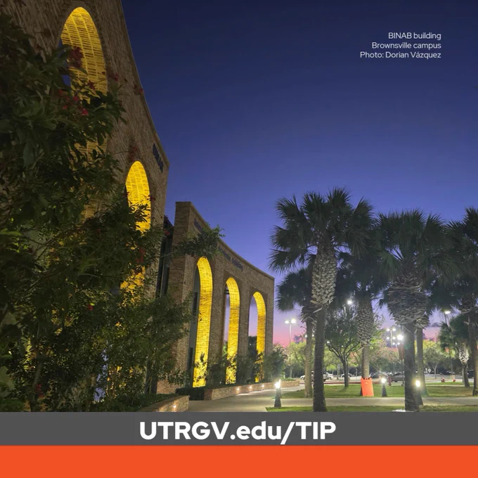 Evening view of UTRGV's BINAB Building on Brownsville Campus. The building's illuminated brick veneer arches curve along a palm-lined walkway beneath a deep blue twilight sky, with hints of pink on the horizon.