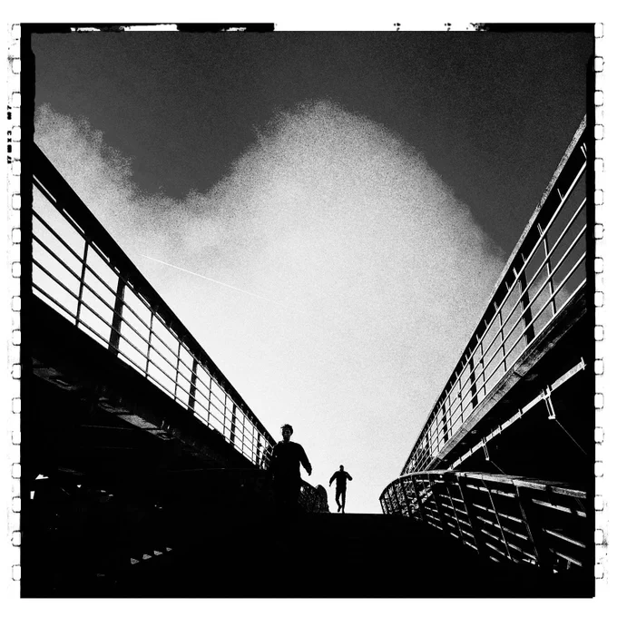 Black-and-white photograph of a pedestrian bridge seen from below, with strong diagonal railings rising on both sides toward a bright sky. Two human silhouettes appear at the top of the stairs, creating a dramatic sense of scale, tension, and movement.
