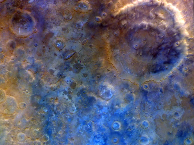 Enhanced-color image of Mars showing a vast, ancient landscape. In the upper right you can see the large double-ring Huygens Crater, a prominent circular formation about 470 kilometers wide with a clear inner ring. Around it, the southern highlands are covered with lots of smaller craters. The whole area shows warm tones of red/orange and brown, with hints of blue and purple that highlight differences in dust, sand, and rock across the rough, ancient surface.