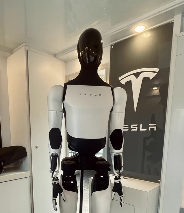 Optimus (Tesla robot) standing inside a white Tesla test cabin with tesla logo in the background.