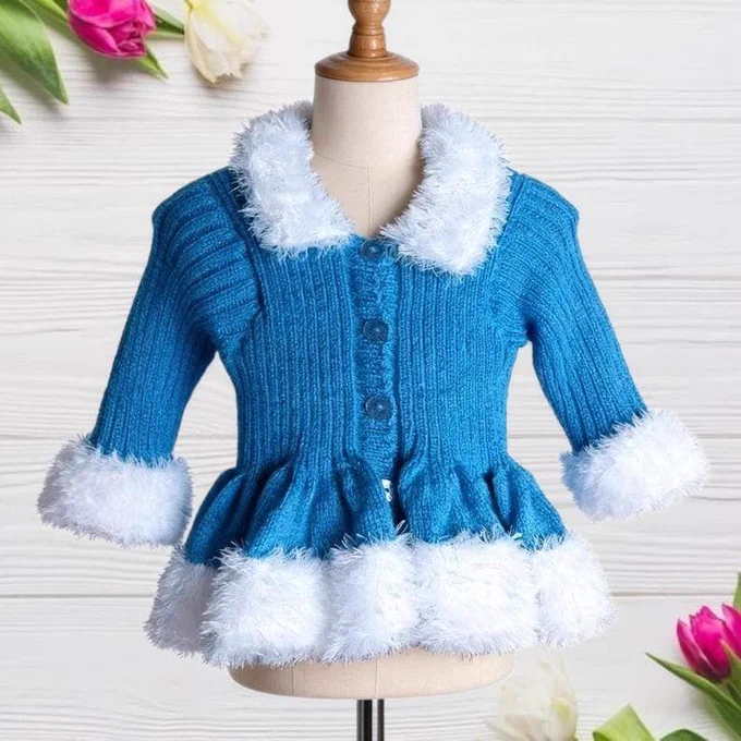 A blue, ribbed-knit toddler cardigan with fluffy white faux-fur trim on the collar, cuffs, and hem sits on a mannequin. It has three blue buttons and a ruffled peplum. The background is white wood with pink and white tulips in the corners.