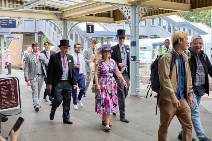 A group of individuals at Ascot station are dressed in both formal attire, such as top hats and suits, and casual clothing. Some are walking, and among them is an individual wearing a purple floral dress with a matching hat. There is a South Western Railway train visible in the background.