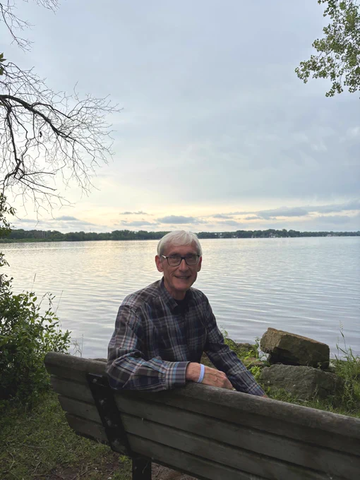 Gov. Evers sits on a bench and smiles at the camera at Lake Kegonsa State Park
