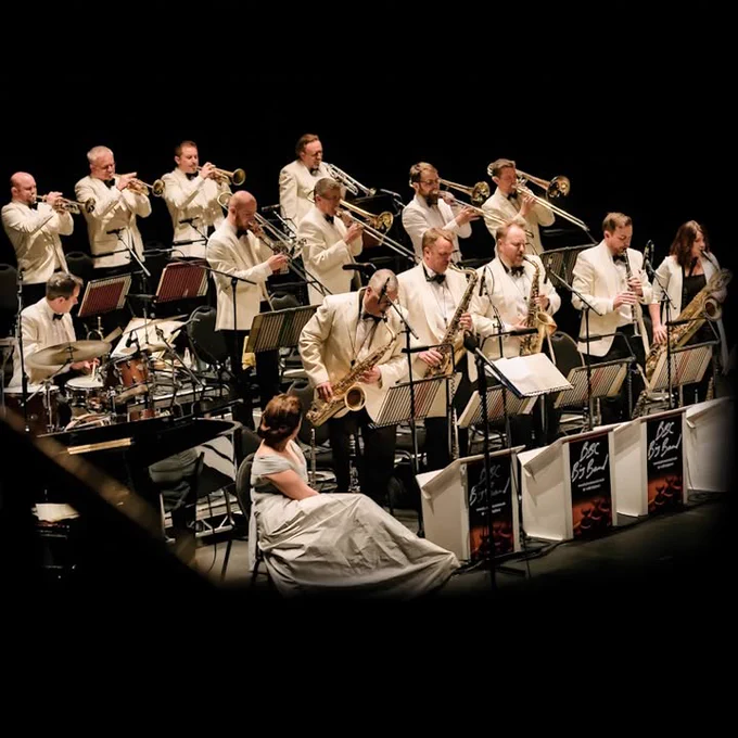 An orchestra in white suits performs on stage with various brass and woodwind instruments, including trumpets and saxophones. The music stands display the name "BBC Big Band." A person sits in front of the ensemble.