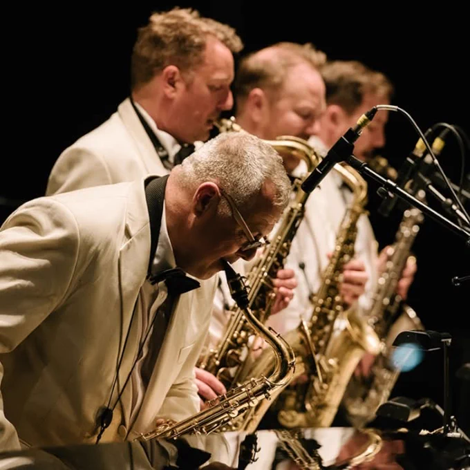 A group of musicians in matching white jackets are playing saxophones on stage. Microphones are positioned in front of them. The background is dark.